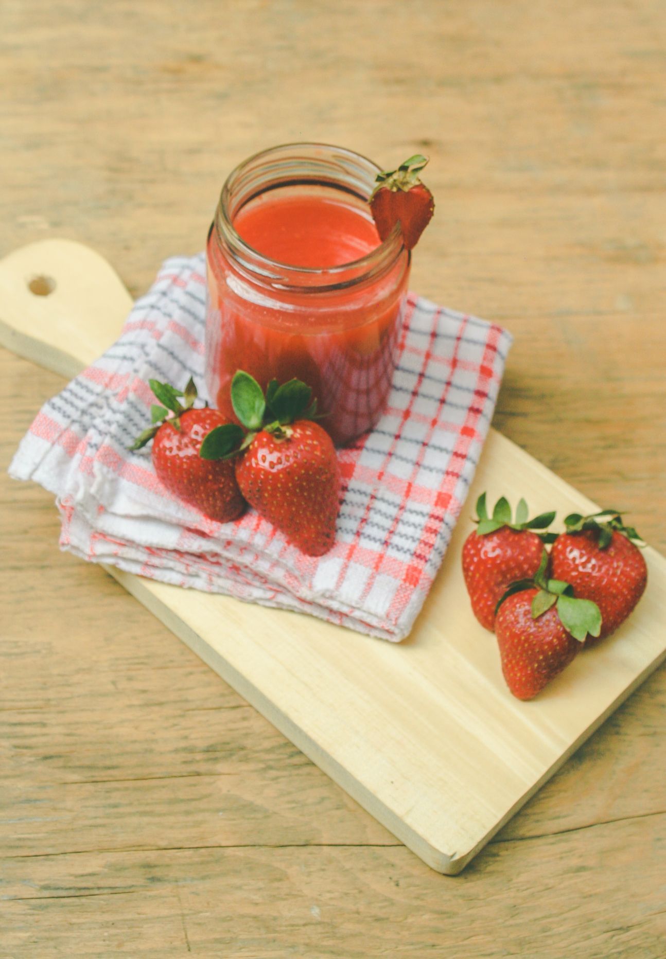 Ein gefülltes Glas mit roter Flüssigkeit und drumherum auf einem Holzbrett einzelne Erdbeeren.
